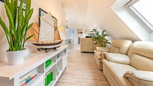 A modern living room with a sofa, a bookshelf, a potted plant, a model sailboat, and a large window.