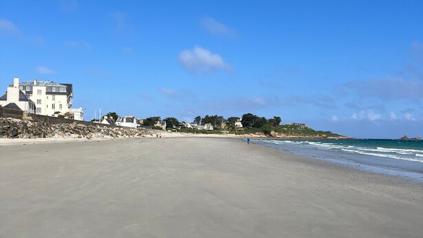 A beach with a large building on the left, a clear sky, and calm sea.