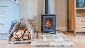A cozy room with a wood-burning stove, a wicker basket filled with firewood, and a wooden cabinet. - Captain's Quarters, a ground floor apartment in Aldeburgh (Aldeburgh)