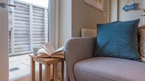 A cozy living room with a grey sofa, a blue pillow, and a wooden side table with an open book. - Captain's Quarters, a ground floor apartment in Aldeburgh (Aldeburgh)