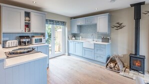 A modern kitchen with light blue cabinets, a central island, a wood-burning stove, and a view of the outdoors through a glass door. - Captain's Quarters, a ground floor apartment in Aldeburgh (Aldeburgh)
