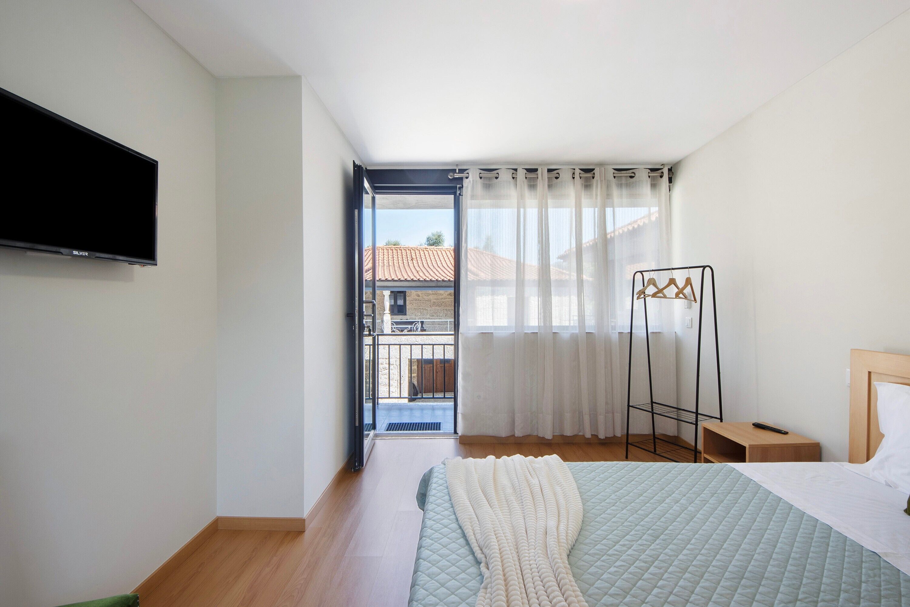 Ein Schlafzimmer mit Bett, Fernseher, Fenster mit Vorhängen, Holzboden und Balkon.