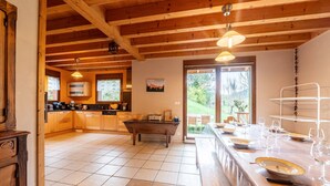 A spacious kitchen with wooden beams, a dining area with a long table set for a meal, and a view of the outdoors through a glass door.