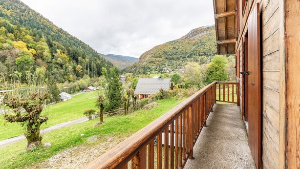 A wooden balcony with a view of a valley, a house in the distance, and a tree with a unique trunk shape.