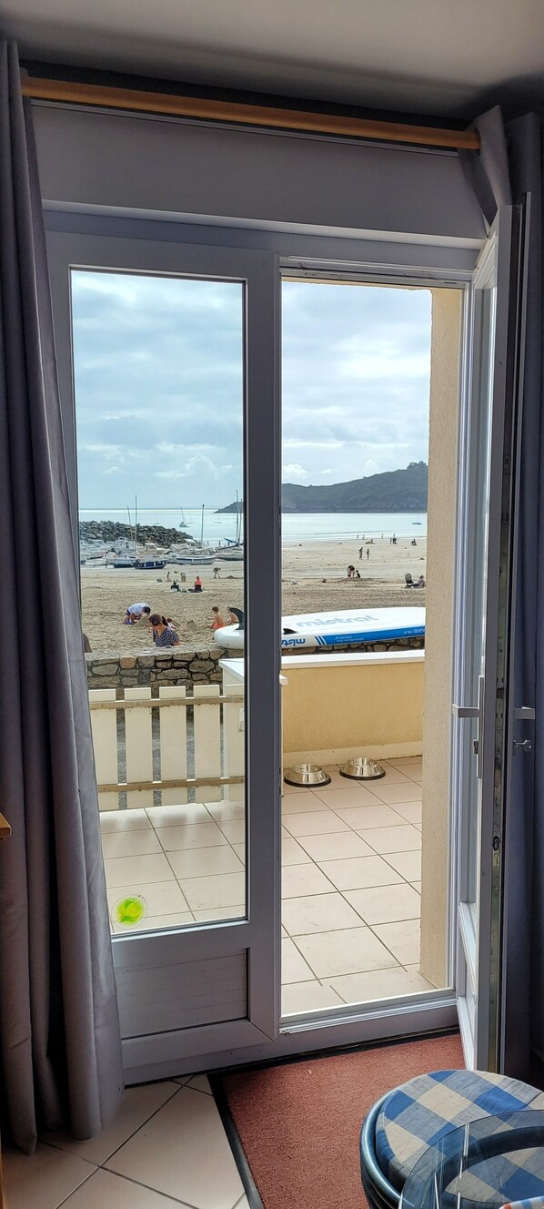 A room with a sliding glass door leading to a balcony with a view of a beach, a pool, and a stone wall.