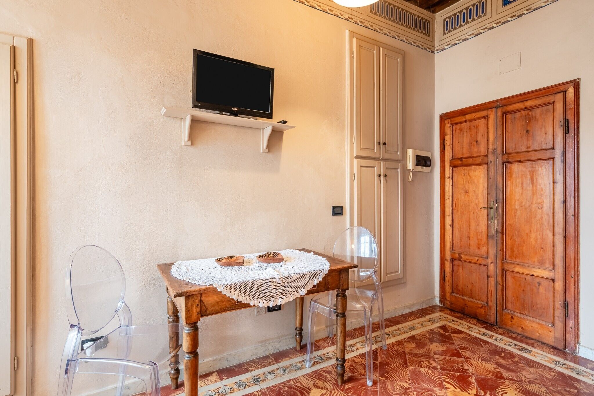 Une pièce avec un téléviseur posé sur une étagère, une table en bois recouverte d’une nappe en dentelle, des chaises transparentes et une grande armoire en bois.