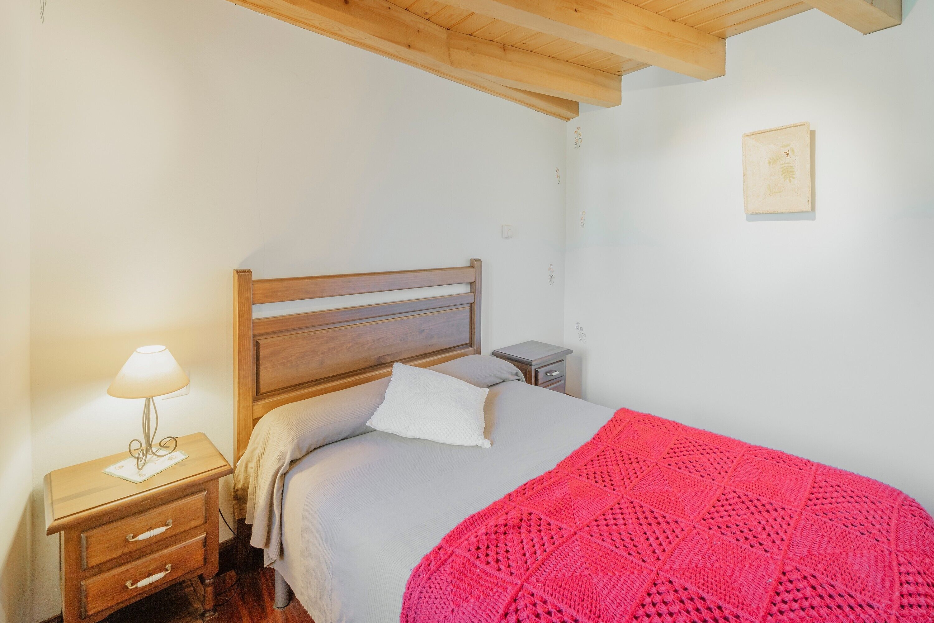 Een slaapkamer met een houten hoofdbord, een nachtkastje met een lamp, en een klein fotokader aan de muur. Er ligt een rood gebreid plaid op het bed.