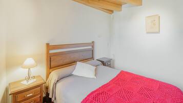 Een slaapkamer met een houten hoofdbord, een nachtkastje met een lamp, en een klein fotokader aan de muur. Er ligt een rood gebreid plaid op het bed.