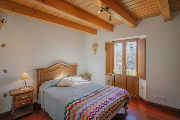 A bedroom with a wooden bed, a nightstand, a lamp, a window with a view, and a wooden ceiling.