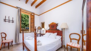 Ein Schlafzimmer mit einem hölzernen Himmelbett, zwei Stühlen, einem Nachttisch, einer Lampe und einem Fenster mit Vorhängen.