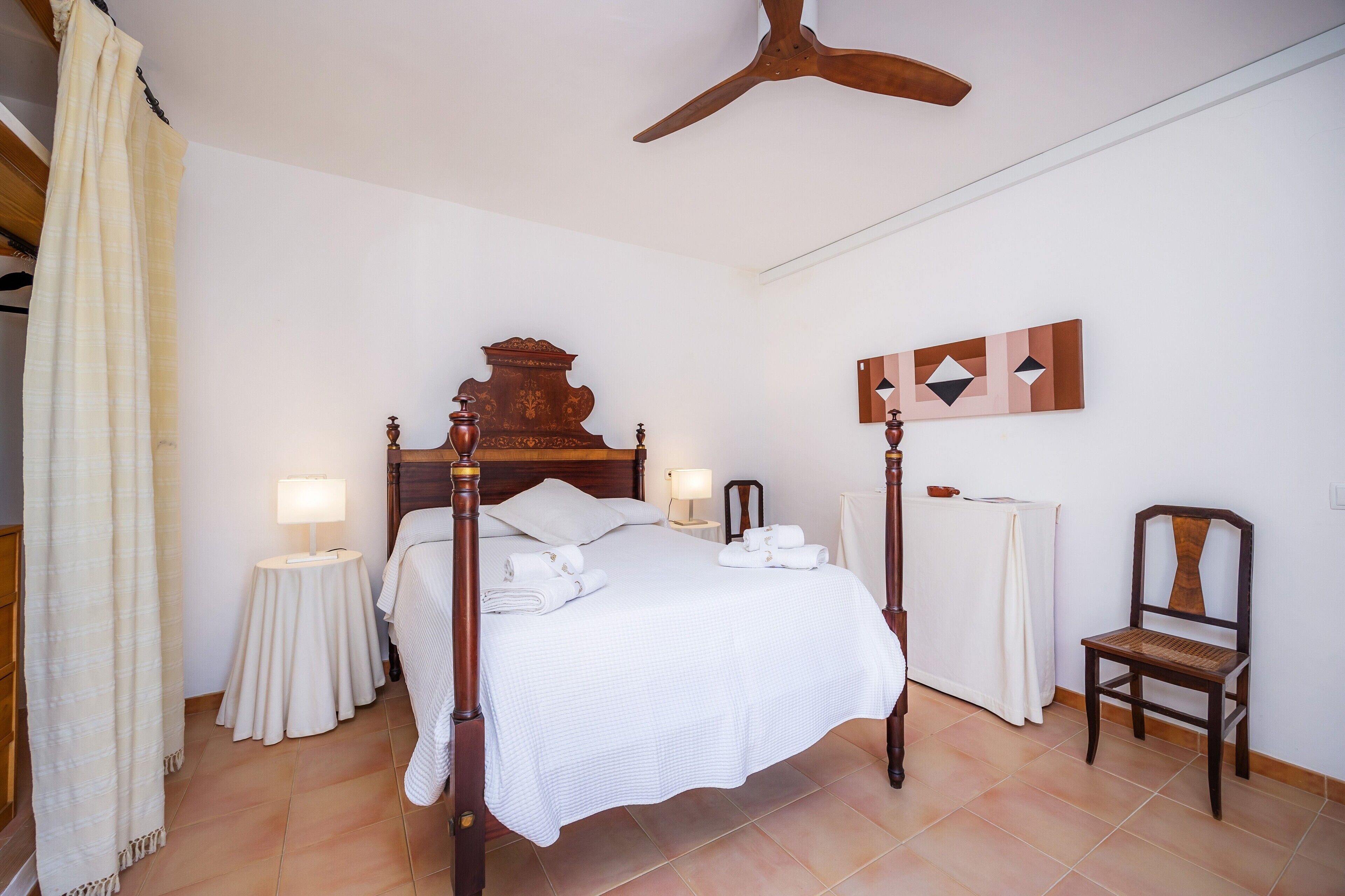 A neatly made bed with pillows and a folded towel, a bedside table with a lamp, a wooden chair, and a ceiling fan.