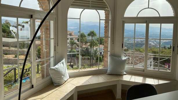 Una habitación con amplios ventanales arqueados, un escritorio blanco y vistas a un paisaje montañoso.