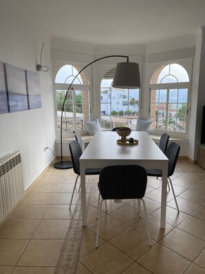 A dining area with a white table, black chairs, a floor lamp, and a view through large arched windows.