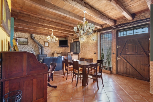 A rustic interior with exposed wooden beams, a stone wall, a dining area with a wooden table and chairs, a kitchen with a stove, and a large wooden door.