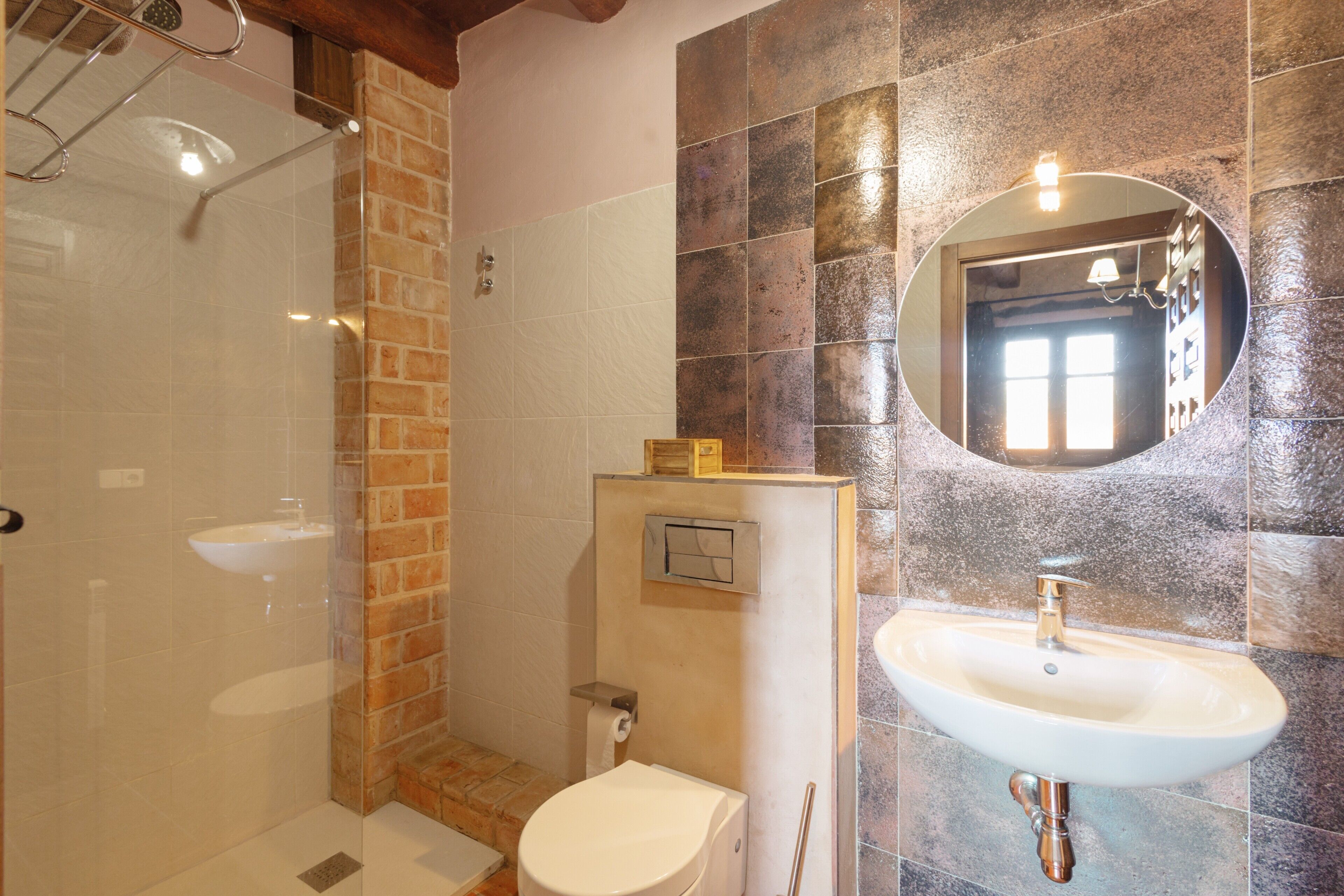 Un bany amb una cabina de dutxa de vidre, un vàter blanc, un lavabo amb mirall i una paret combinada amb rajoles de maó i pedra.