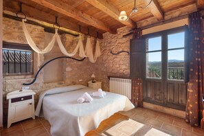 Un dormitorio con cama, mesita de noche, ventana con cortinas, vista al exterior y pared de piedra.