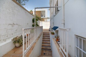 Un étroit couloir extérieur avec une rambarde blanche, des marches carrelées et des plantes en pot.