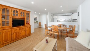 A living room with a TV, wooden cabinets, a dining area with chairs, and a kitchen in the background. - Tomasol Vacation Apartment with Sea Views, Private Terrace, and Wi-Fi (Los Cristianos)