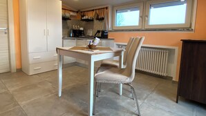 A compact kitchen with white cabinets, a dining table, and a chair. There is a window with blinds, a radiator, and a small countertop with kitchen items. - Villa Giulia - Marguareis (Piemonte)