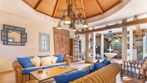 Amplia sala de estar con techo de madera, muebles de mimbre, una mesa central y una estantería.