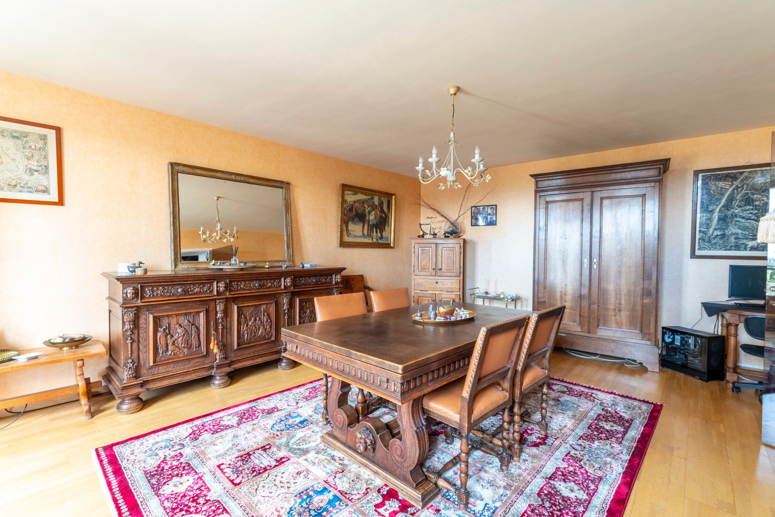 Une salle à manger avec une table en bois, des chaises, un lustre, un grand miroir et un tapis aux motifs rouges et bleus.