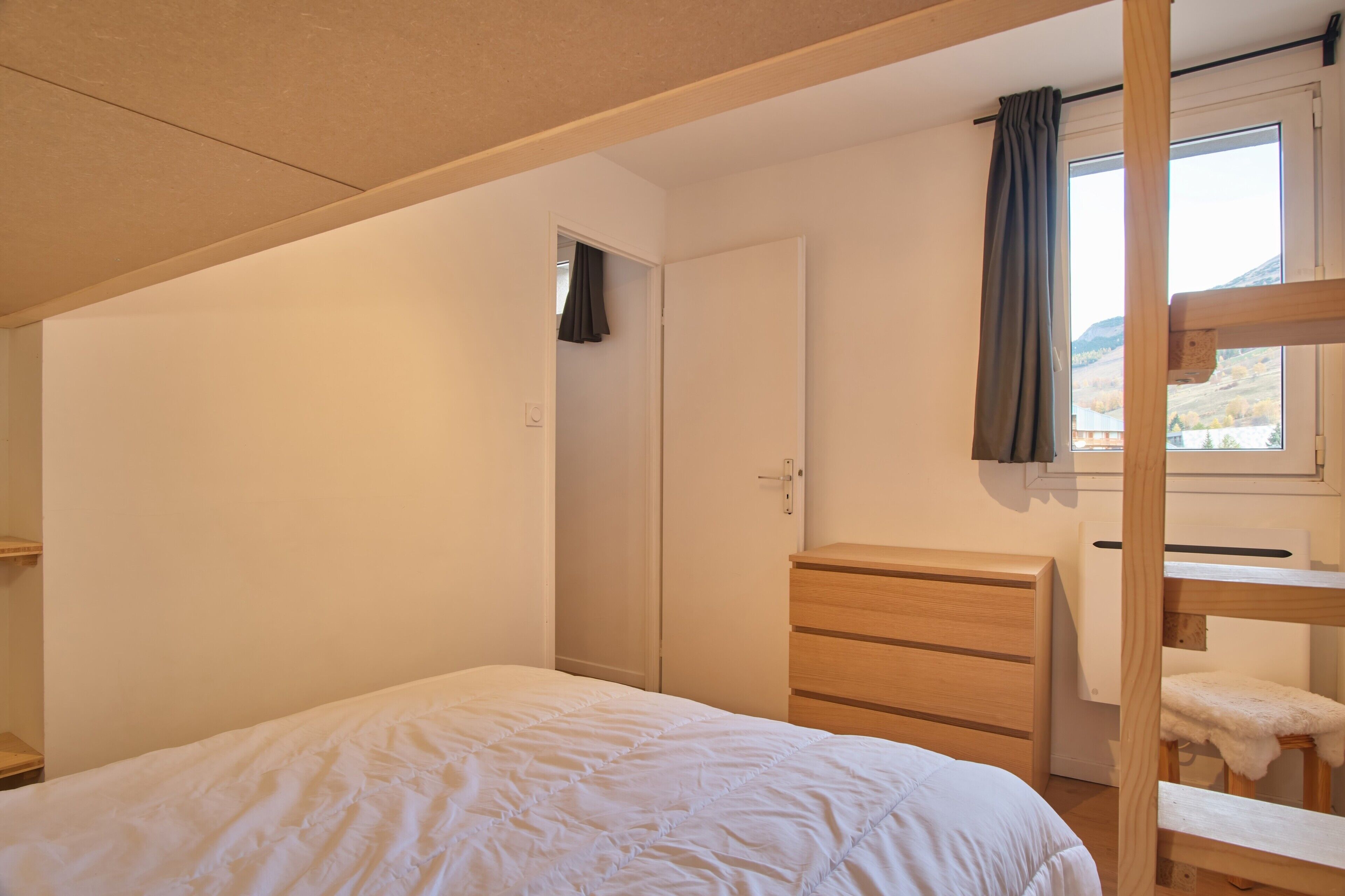 A bedroom with a bunk bed, a wooden dresser, a wardrobe, and a window with a view of a mountain.