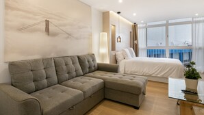 Ein modernes Hotelzimmer mit Ecksofa, einem Bett, einem Glastisch und einem großen Fenster mit Vorhängen.