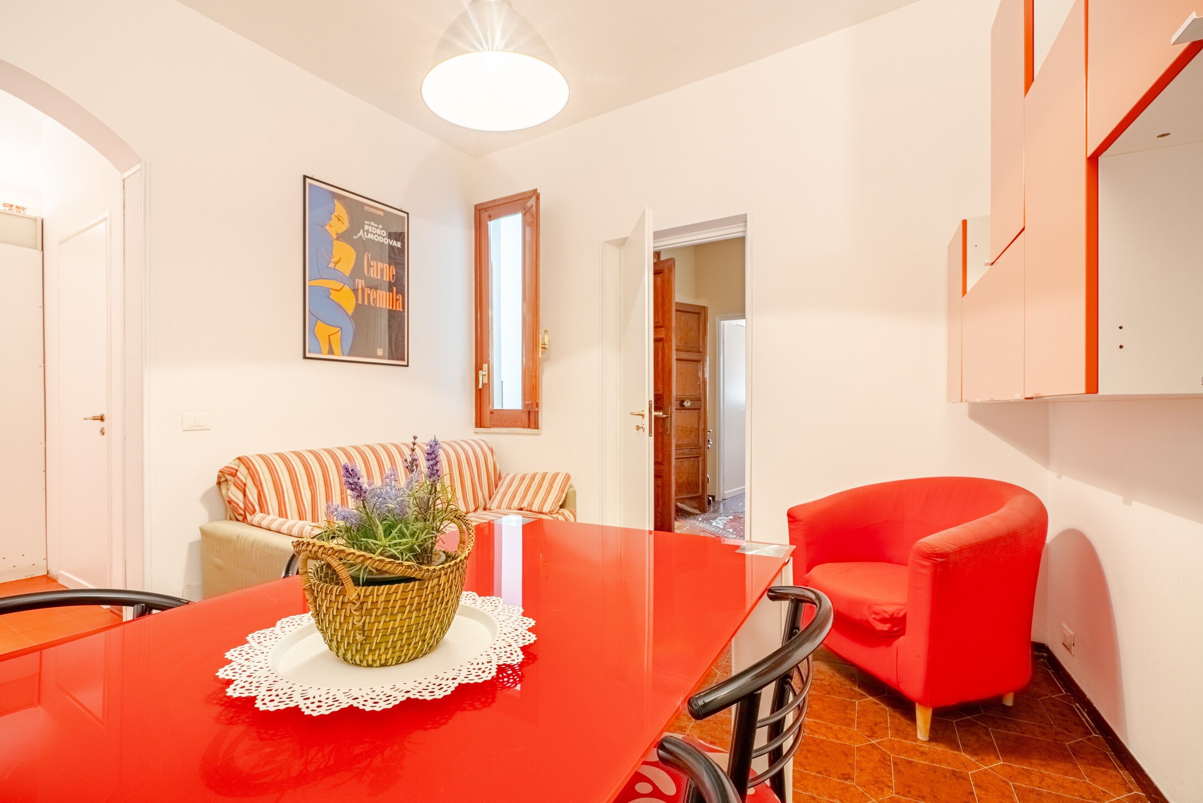 A dining area with a red table, a wicker basket with flowers, a red armchair, and a striped sofa.