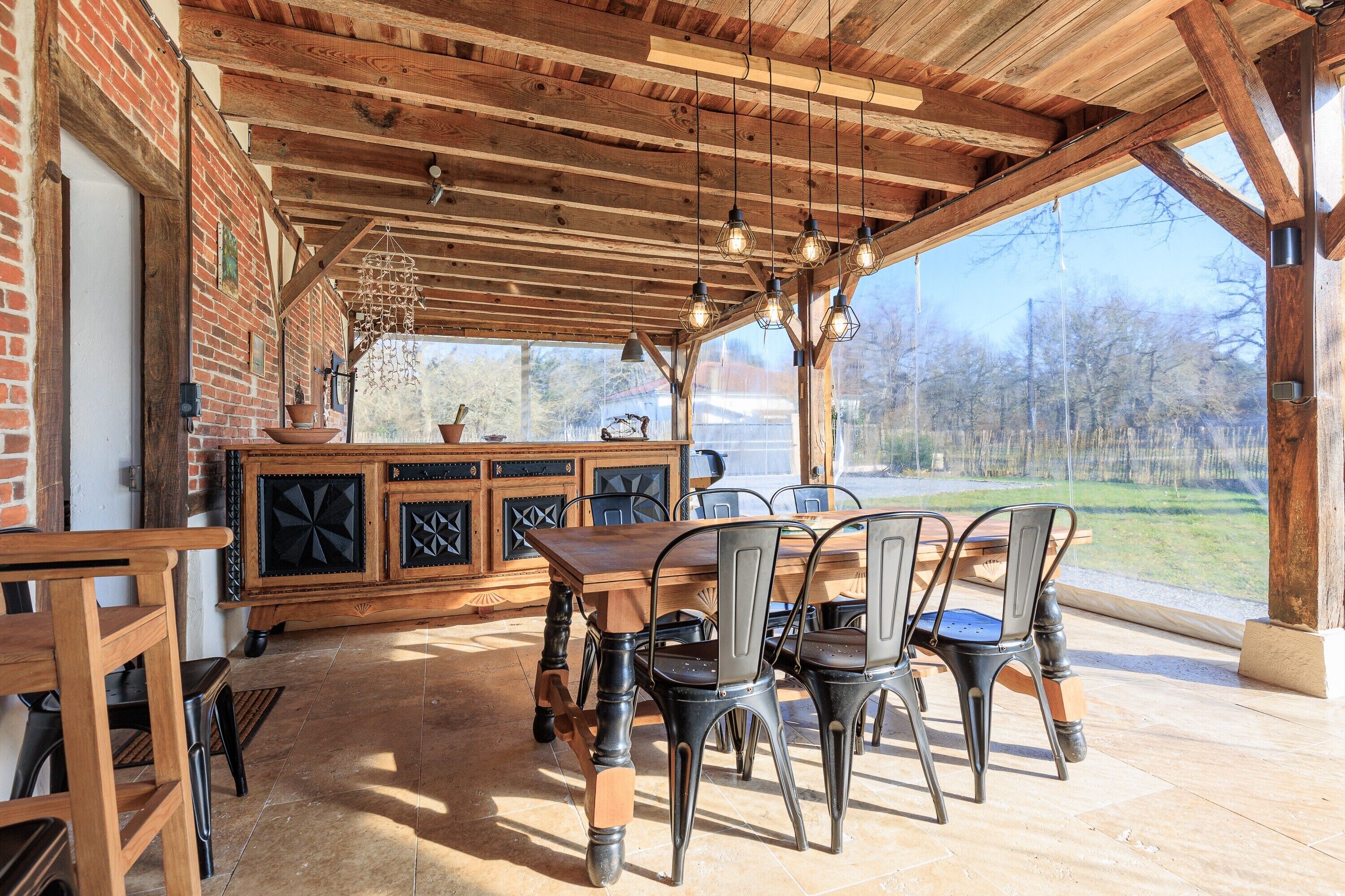 Un espace extérieur abrité, aménagé pour les repas, avec un mur en briques, un plafond en bois et des luminaires suspendus.