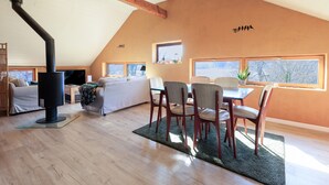 Un loft espacioso con techo alto, vigas de madera a la vista, chimenea, zona de comedor con sillas y mesa, y ventanas grandes que permiten la entrada de luz natural.