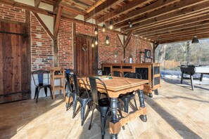Ein rustikaler Essbereich mit Backsteinwand, Holzbalken und einem großen Holztisch, um den herum Metallstühle stehen.