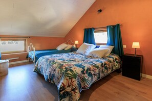 Ein Schlafzimmer mit schrägen Wänden, einem Bett mit floralem Überwurf, zwei Nachttischen mit Lampen und einem Fenster mit Vorhängen.
