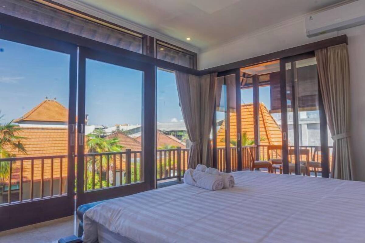 A hotel room with a large bed, a balcony with a view of buildings and palm trees, and a window with curtains.