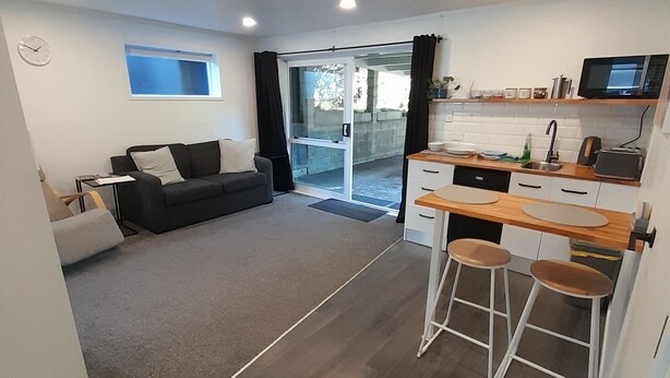 A modern living room with a sofa, a dining area with a kitchen counter, and a large sliding glass door leading to an outdoor area.