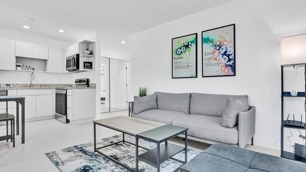 A modern living room with a grey sofa, a coffee table, and a rug. There is a kitchen in the background with white cabinets and a microwave. Two framed artworks are on the wall.