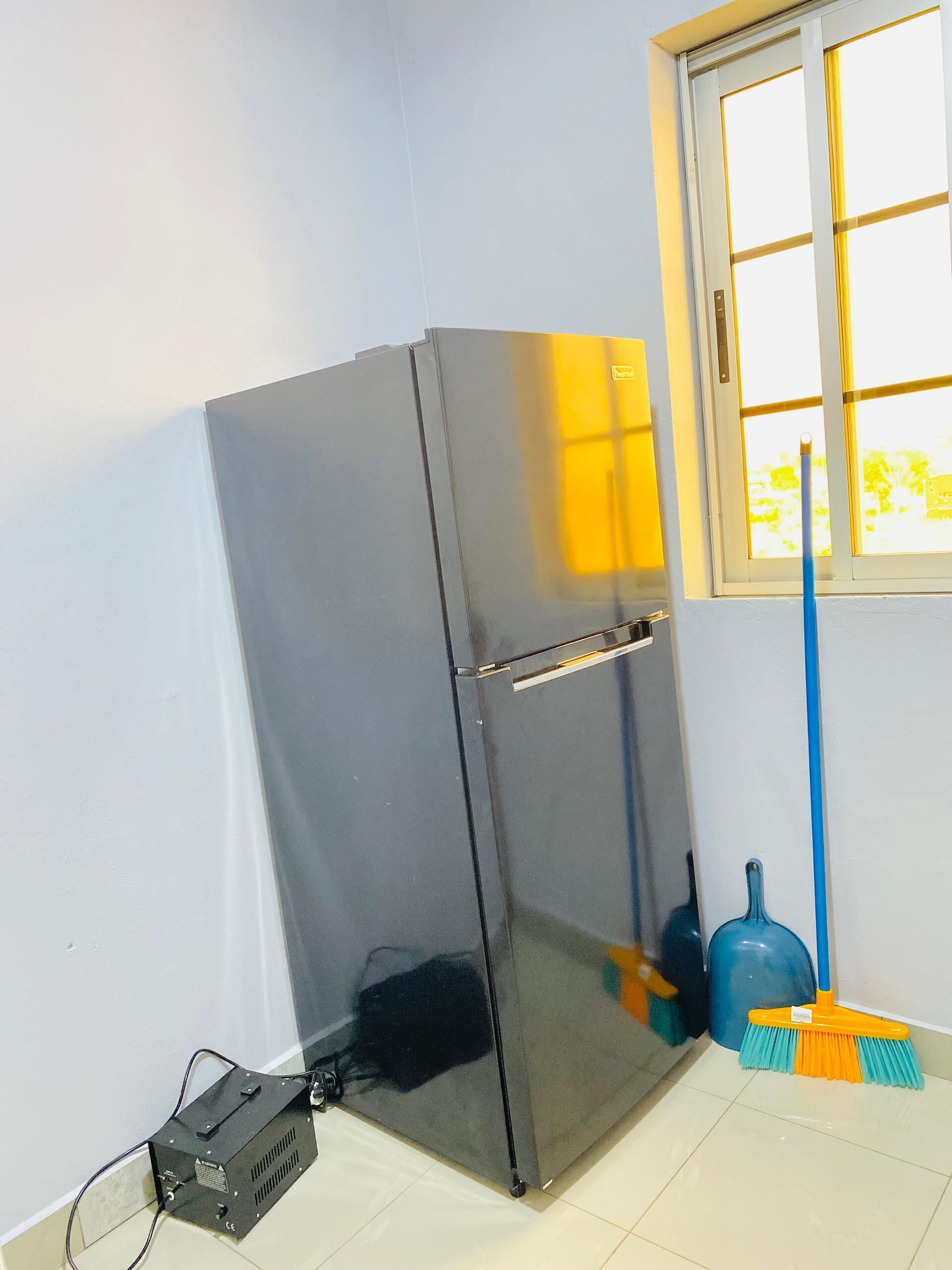 A room with a black refrigerator, a window with a view, a broom, and a power strip.