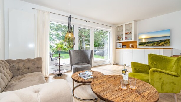 En moderne stue med sofa, lænestol og sofabord. Der er et spiseområde med et bord og stole. Værelset har store vinduer og udsigt til det fri.