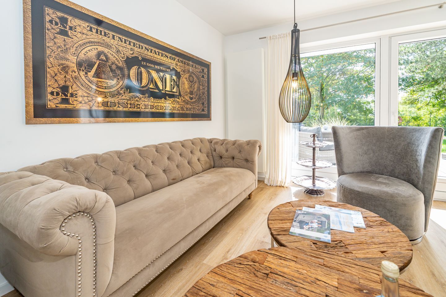 Una sala de estar con un sofá capitonado, una mesa redonda de madera, un sillón gris y una obra de arte grande enmarcada en la pared.