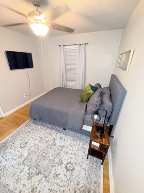 A bedroom with a bed, a ceiling fan, a television mounted on the wall, a window with curtains, and a small wooden side table.