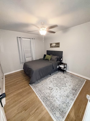 A bedroom with a bed, a nightstand, a ceiling fan, a window with curtains, and a patterned rug.