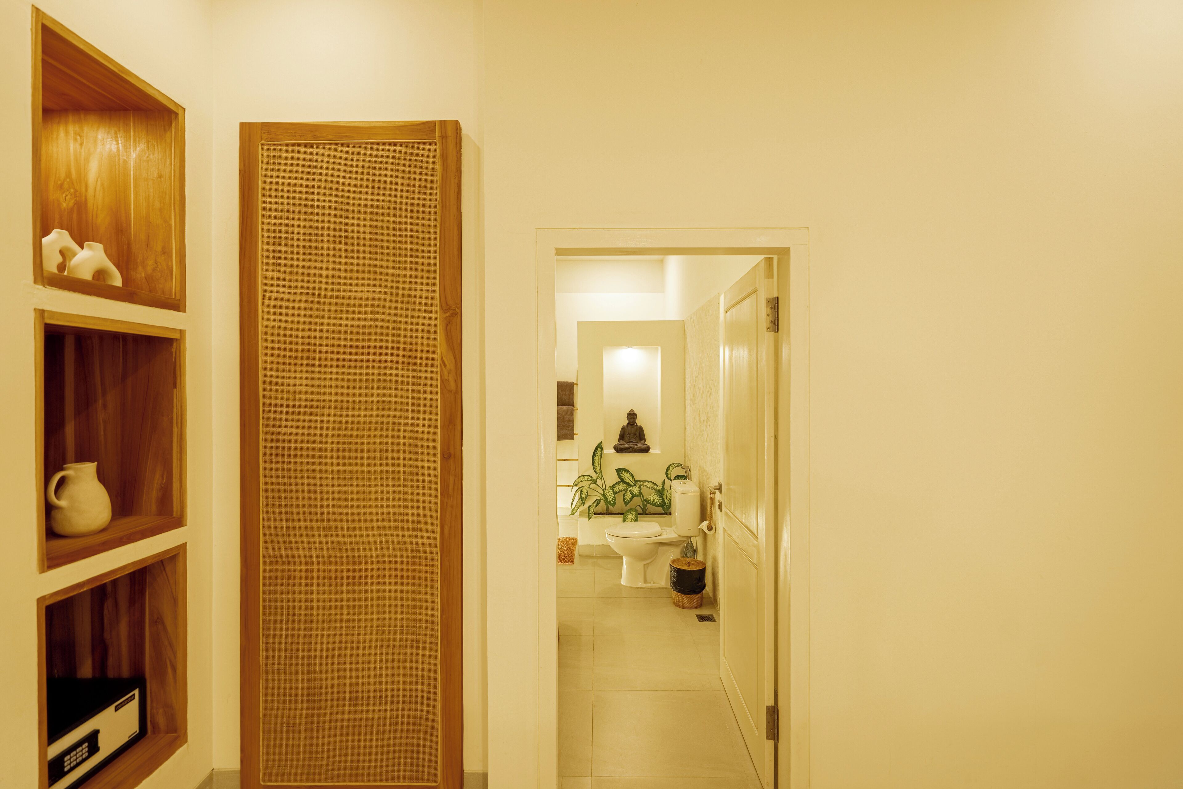 A hallway leading to a bathroom with a toilet, sink, and a potted plant. There are wooden shelves on the left with a vase and a pitcher.