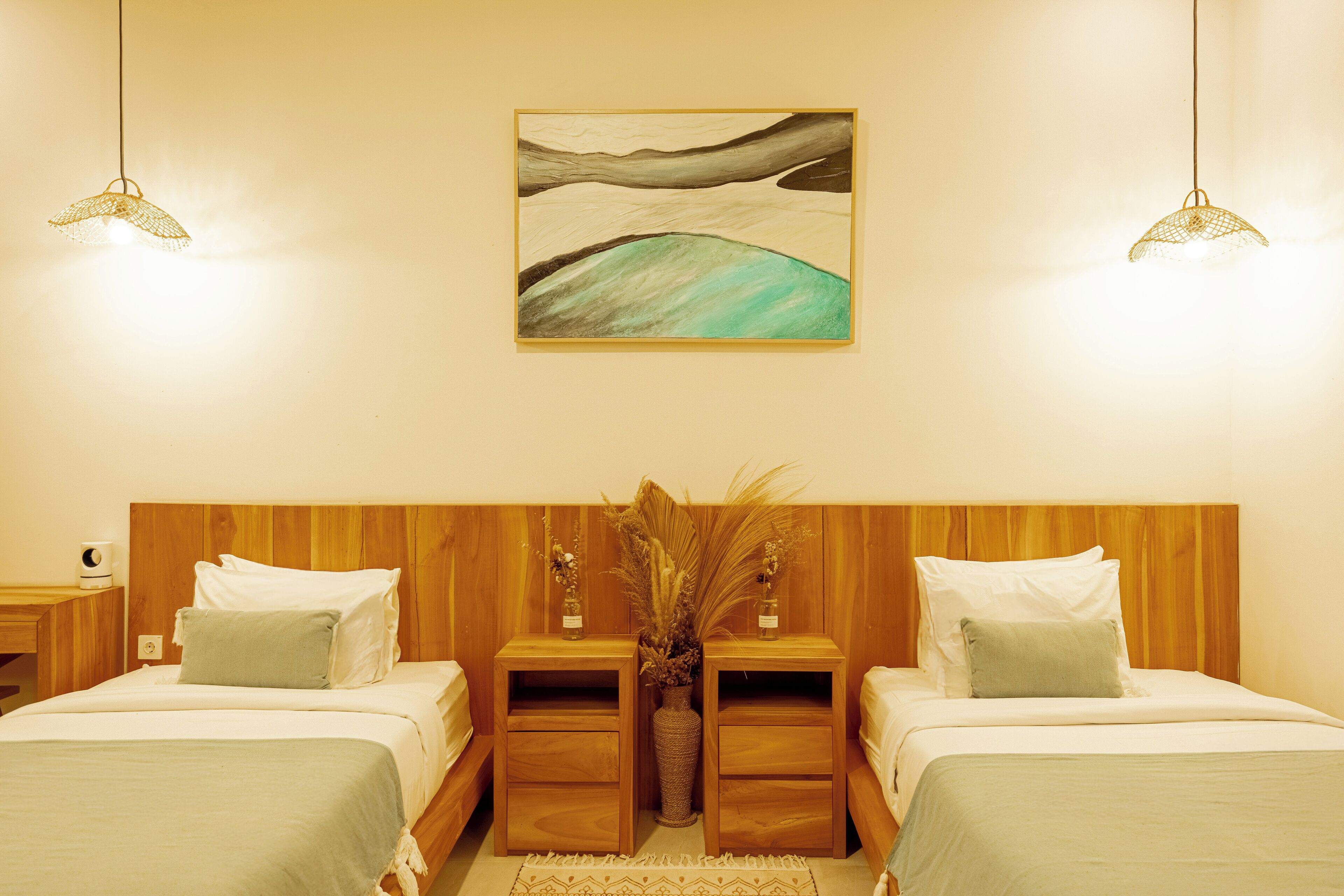 A hotel room with two beds, wooden headboards, bedside tables, a vase with dried plants, and a painting on the wall.