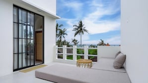 Ein Balkon mit weißer Wand, einer Glastür, einer gepolsterten Sitzecke und Blick auf Palmen und einen klaren Himmel.
