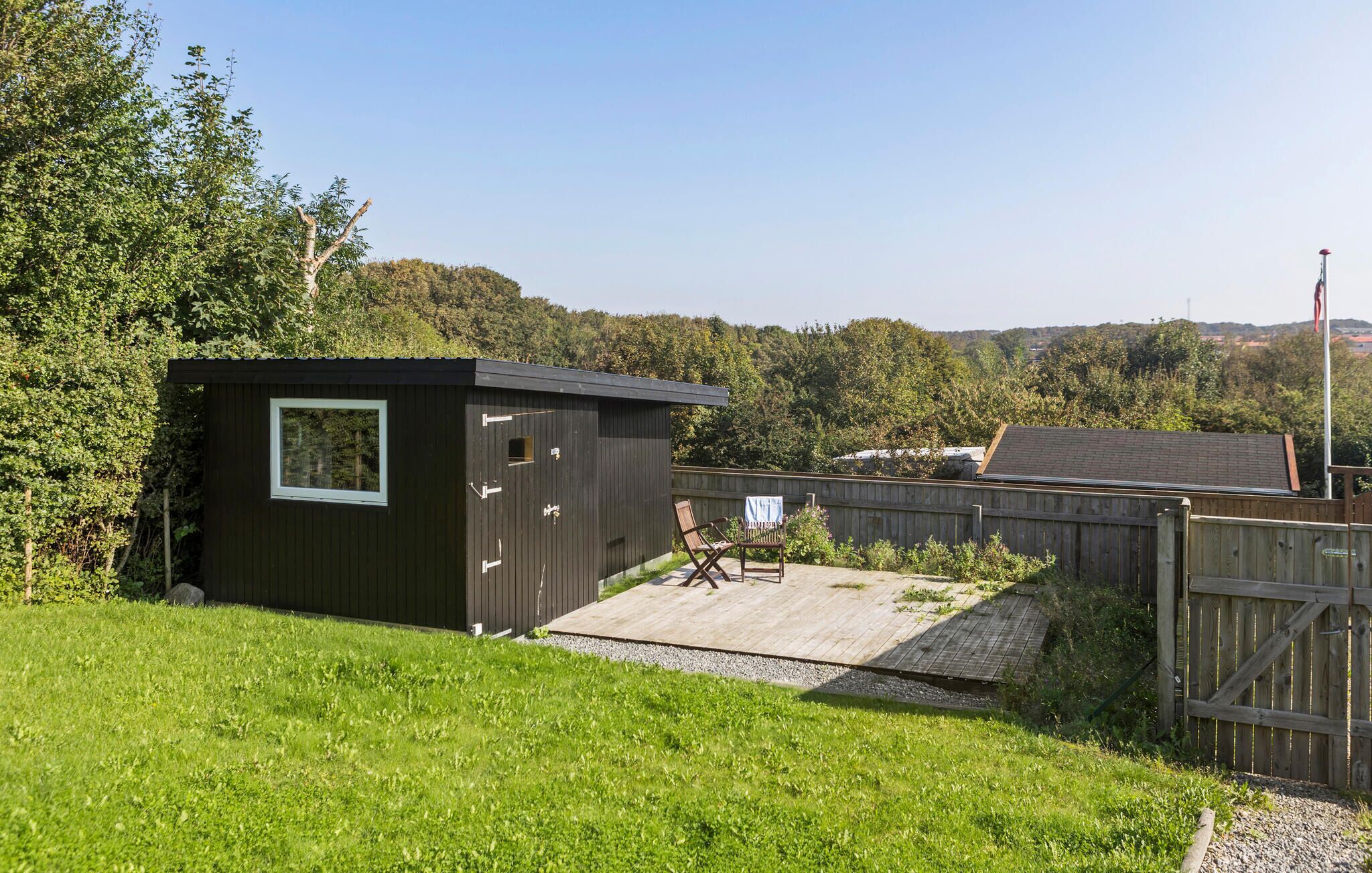 Un petit abri en bois avec une fenêtre, une terrasse en bois et un espace clôturé avec une chaise.