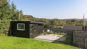 Un petit abri en bois avec une fenêtre, une terrasse en bois et un espace clôturé avec une chaise.