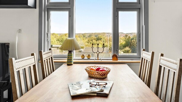 Une table à manger avec un journal, des verres et un panier de fruits, placée près d’une fenêtre donnant sur des arbres et des collines.