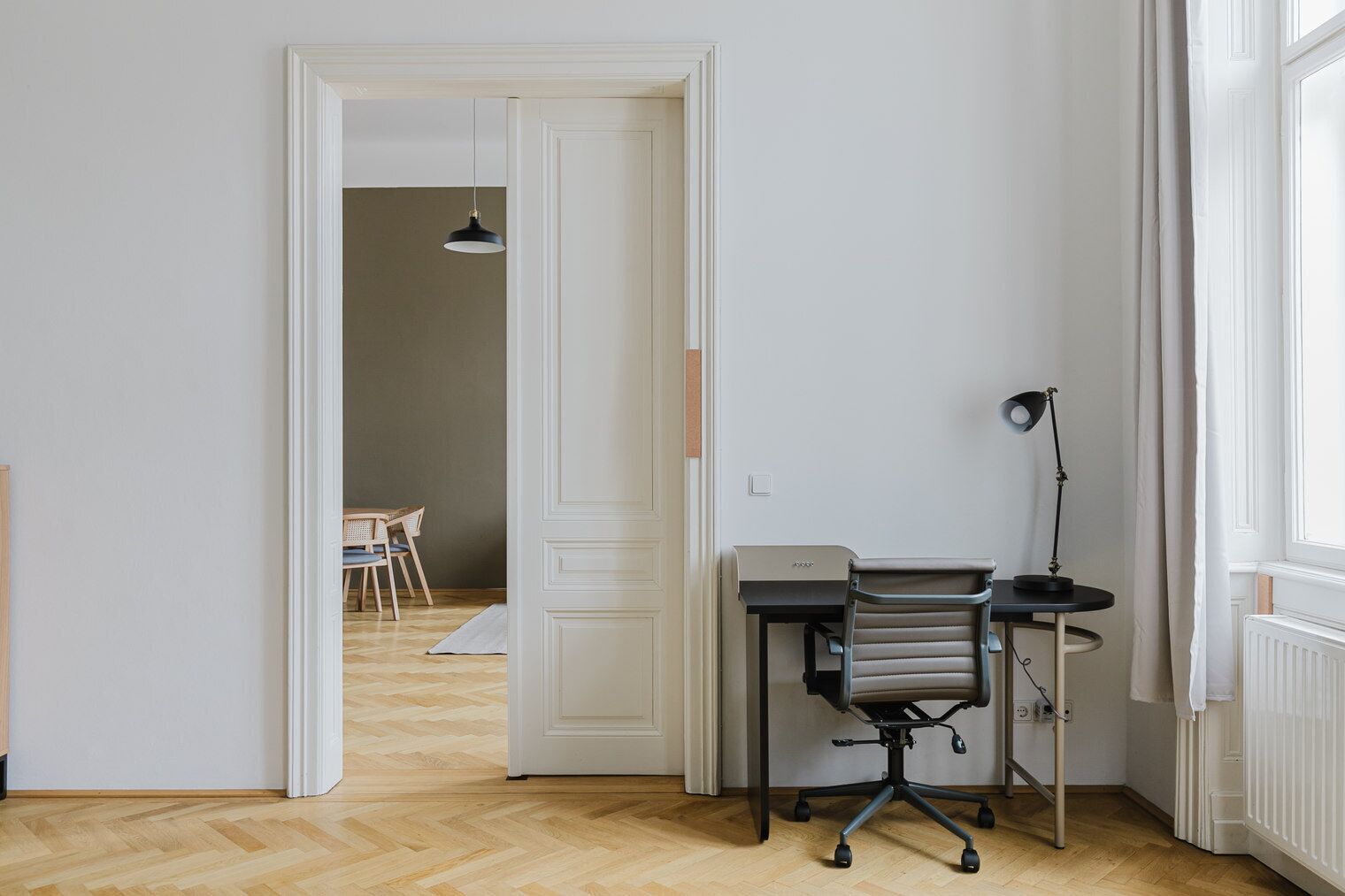 Une pièce comprenant un bureau, une chaise, une lampe et donnant sur une autre pièce où se trouvent une table et des chaises.