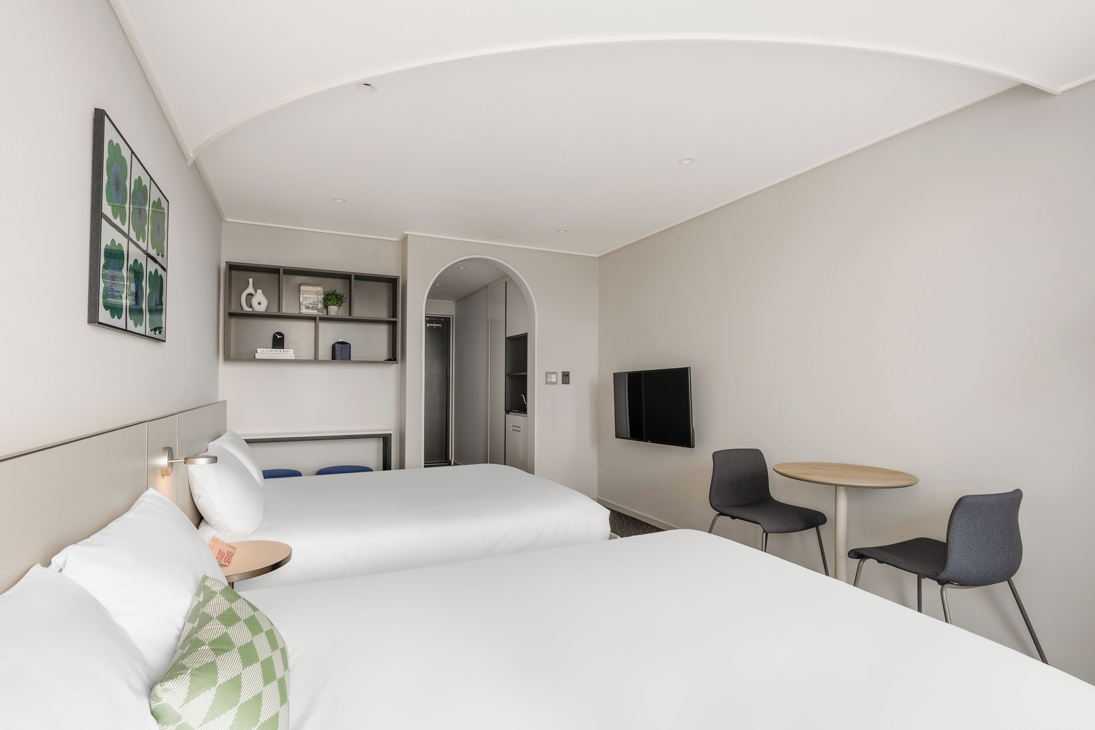 A modern hotel room with two beds, a small table, two chairs, a TV, and a bookshelf.