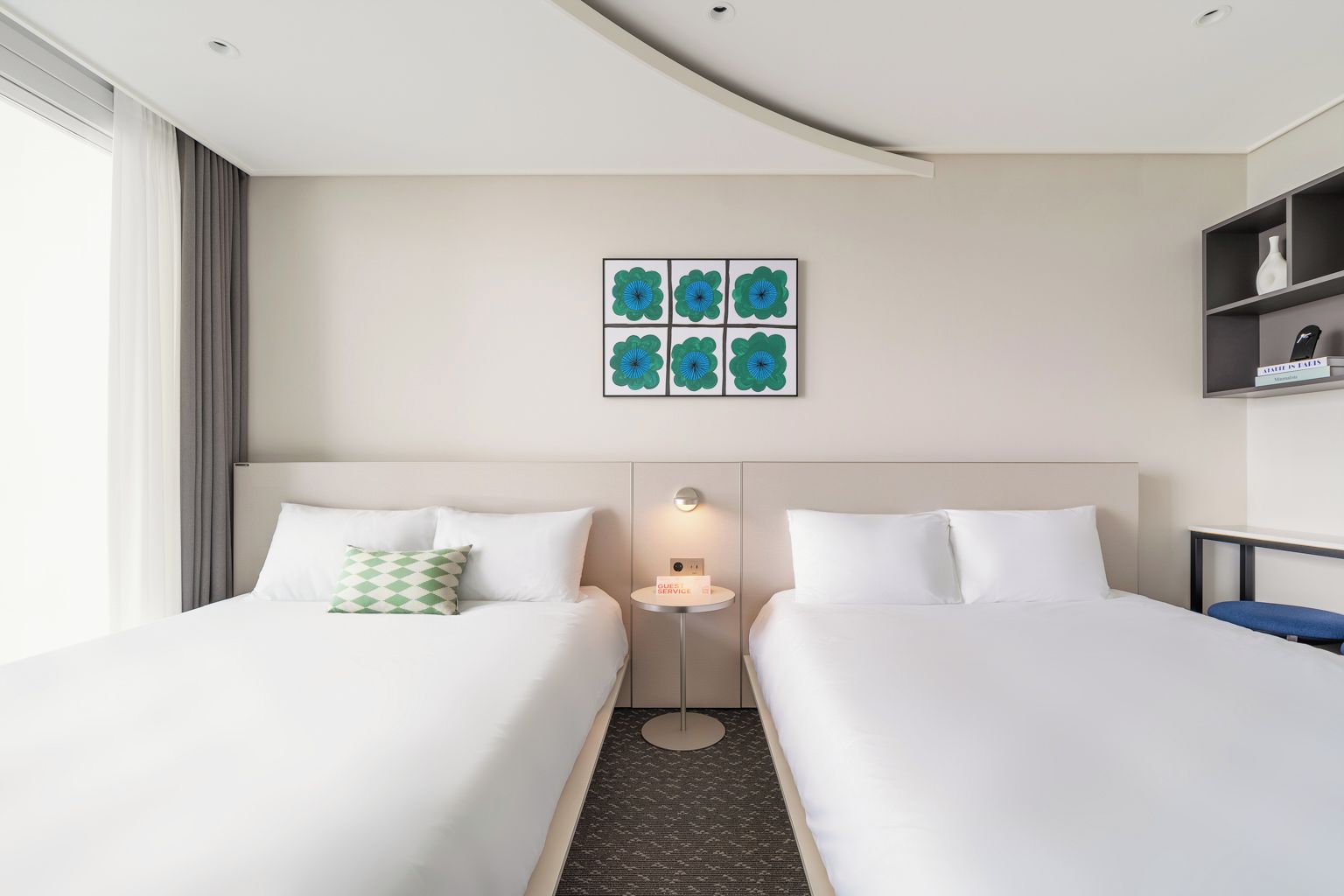 Quarto de hotel com duas camas, uma mesinha de cabeceira com abajur, uma obra de arte na parede e uma estante com um vaso.