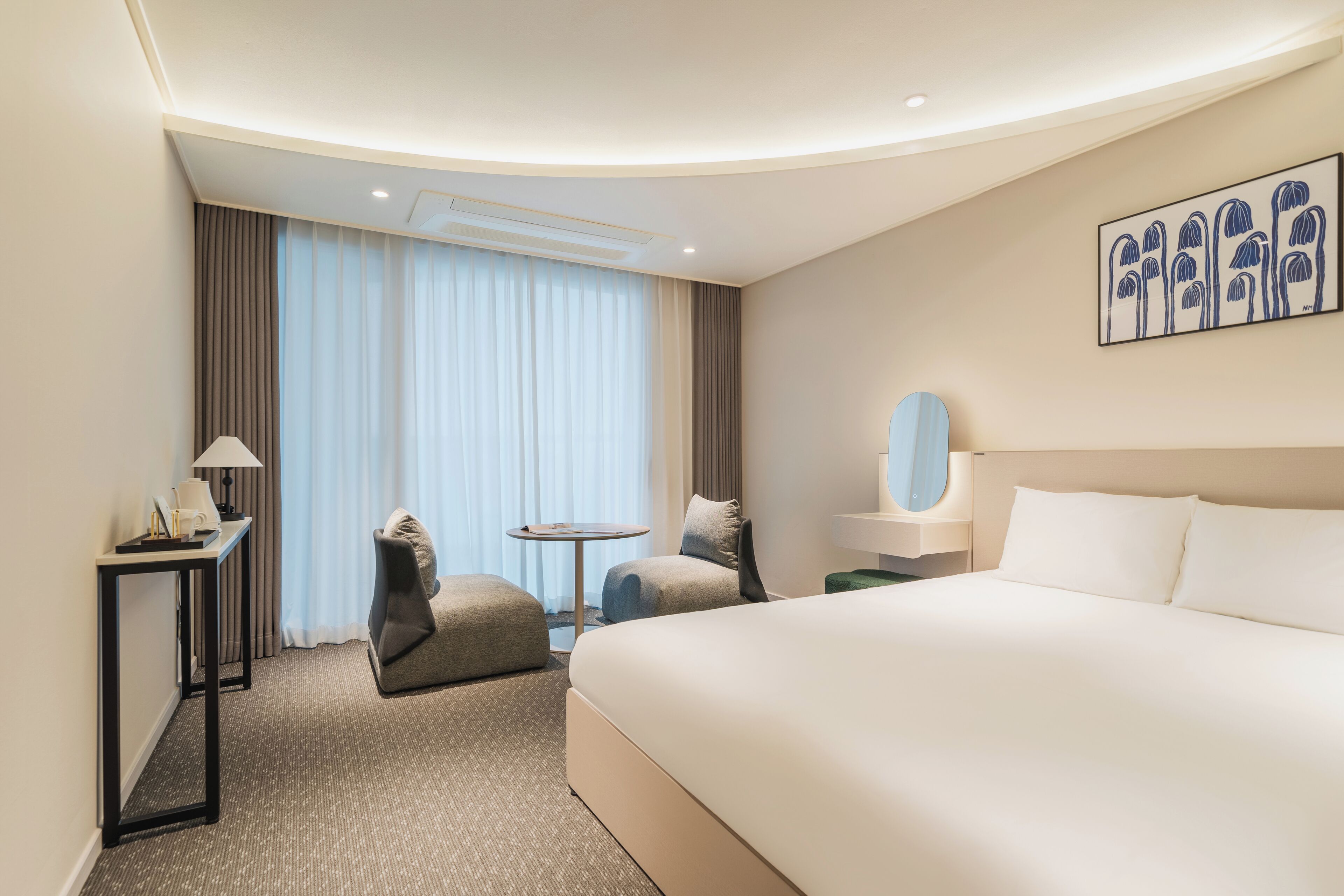 Una habitación de hotel moderna con una cama grande, una mesita redonda pequeña, dos sillas, un escritorio con lámpara y un ventanal con cortinas.